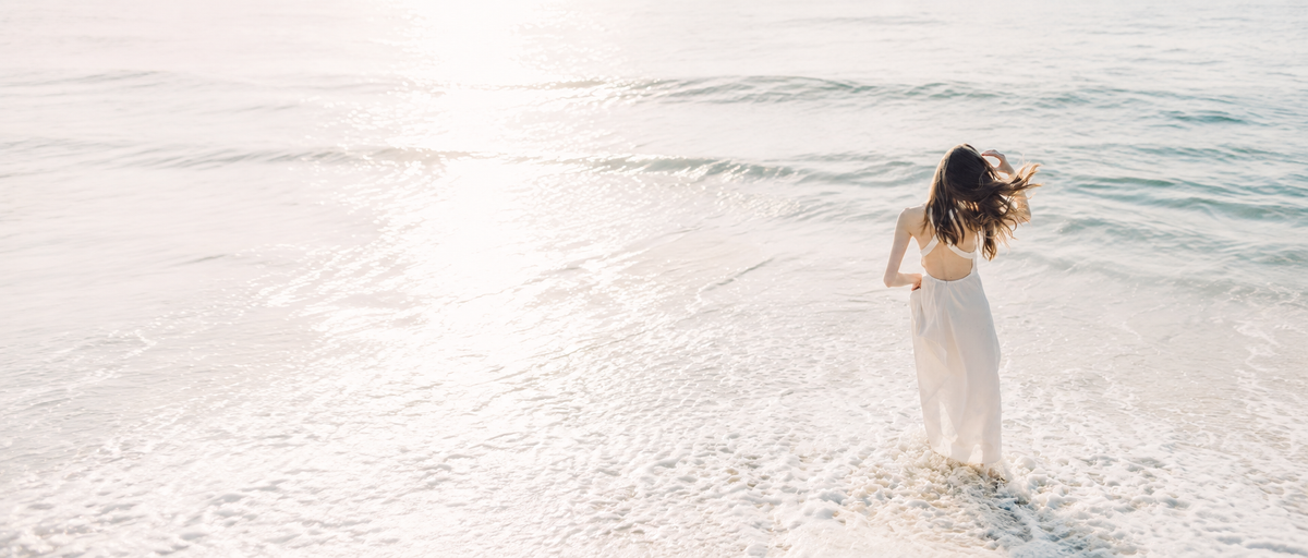 A quiet moment by the sea, with a woman in a white dress enjoying the sunlit waves.